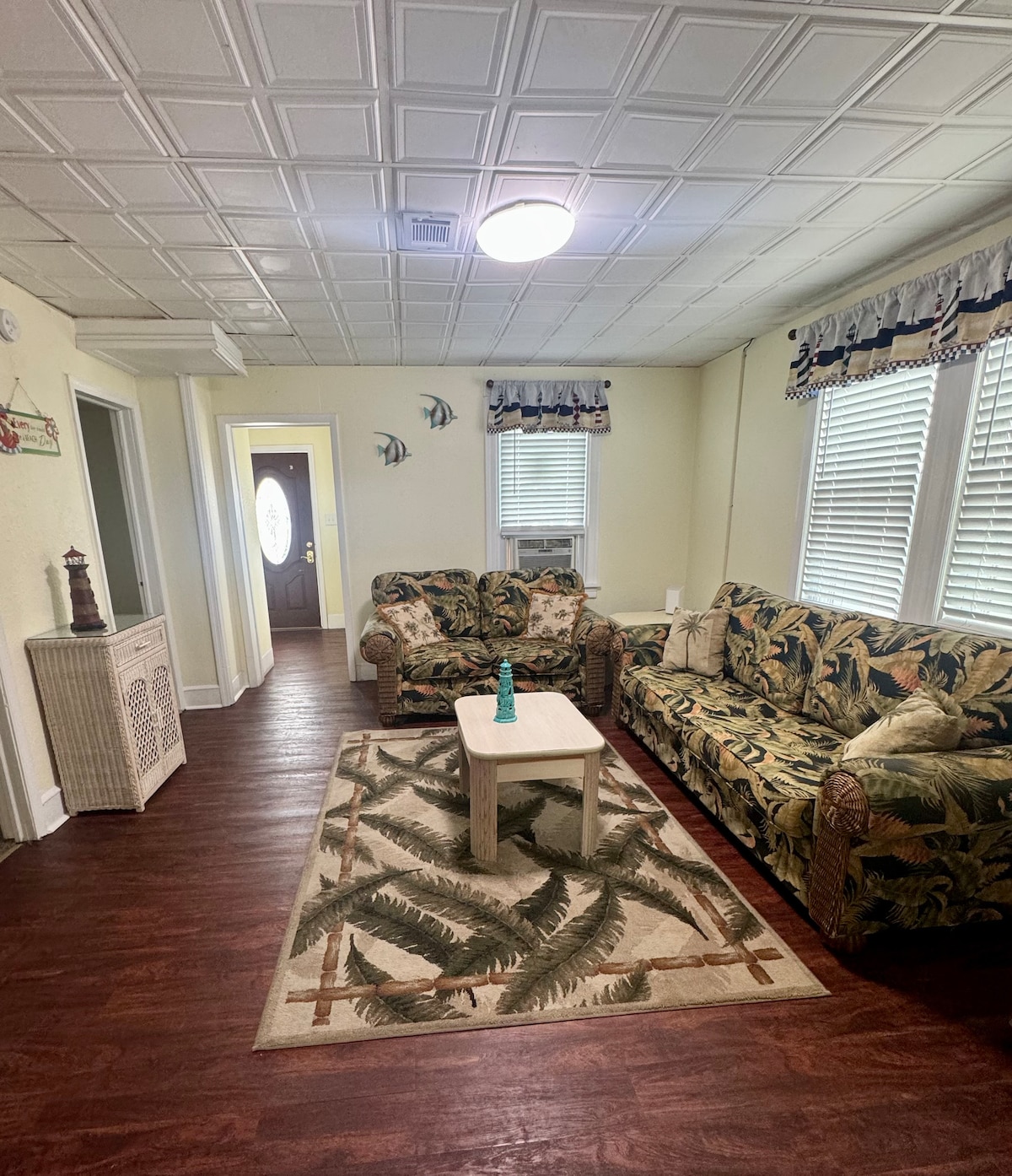 A cozy living area features two patterned sofas and a small coffee table placed on a rug with a leaf design. Natural light fills the space through multiple windows, and wall-mounted decor adds subtle charm. A doorway leads to another room.