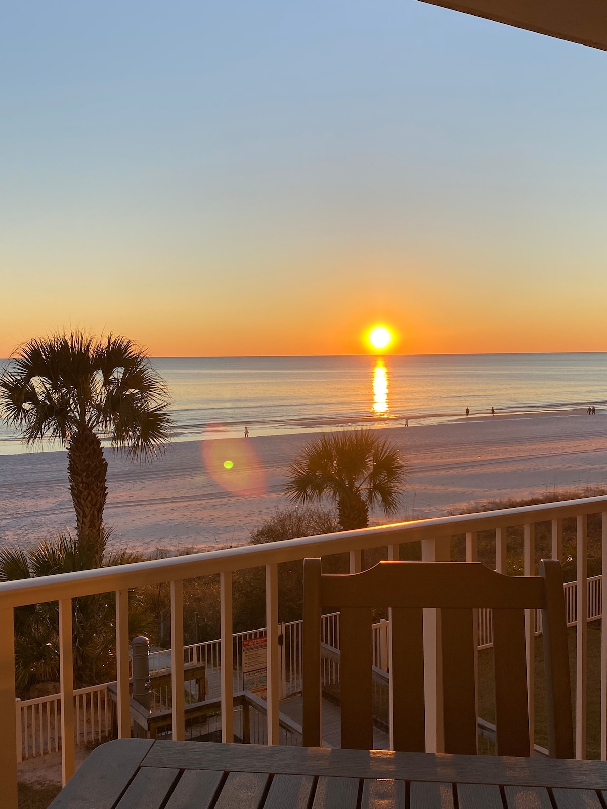 A stunning view features a vibrant sunset over the ocean, with the sun descending towards the horizon. Silhouettes of palm trees are visible in the foreground, enhancing the coastal scenery. The beach is lined with gentle waves and distant figures strolling along the shore.