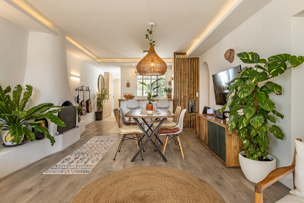 A modern dining area is situated under warm lighting, featuring a round table surrounded by four chairs. Lush green plants are positioned throughout the space, adding a natural touch. A decorative rug lies beneath the table, and a cozy seating area is visible in the background.