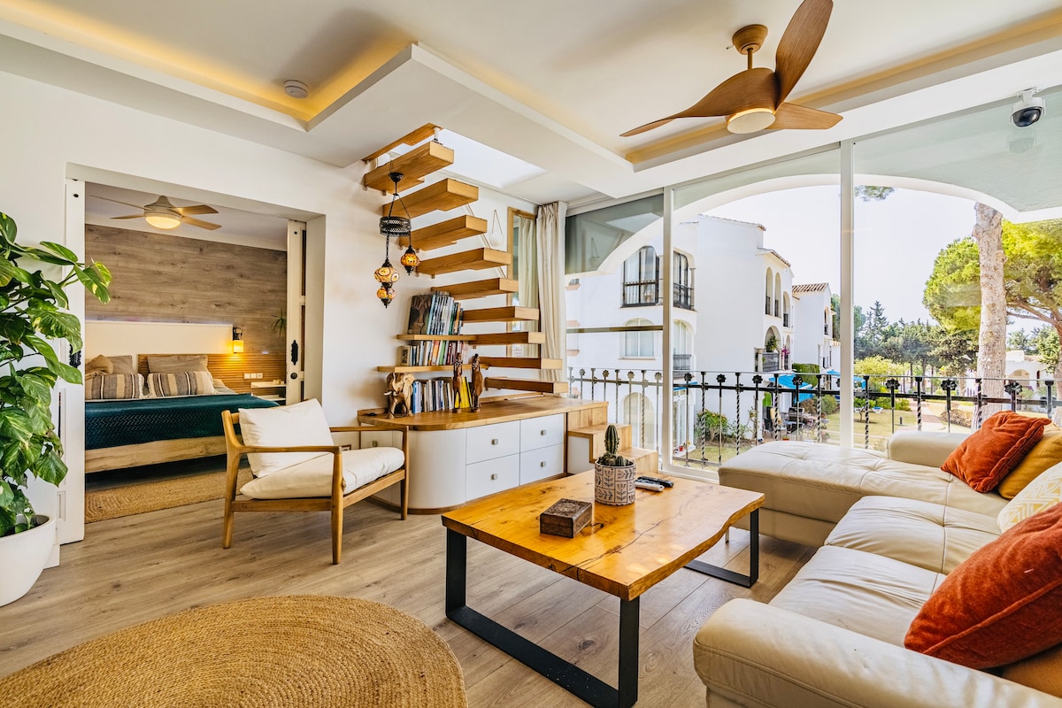 A spacious living area is highlighted by a light-colored sectional sofa and a wooden coffee table. A wooden staircase with shelving leads to a sleeping area. Large glass doors invite natural light and showcase views of the surrounding gardens.