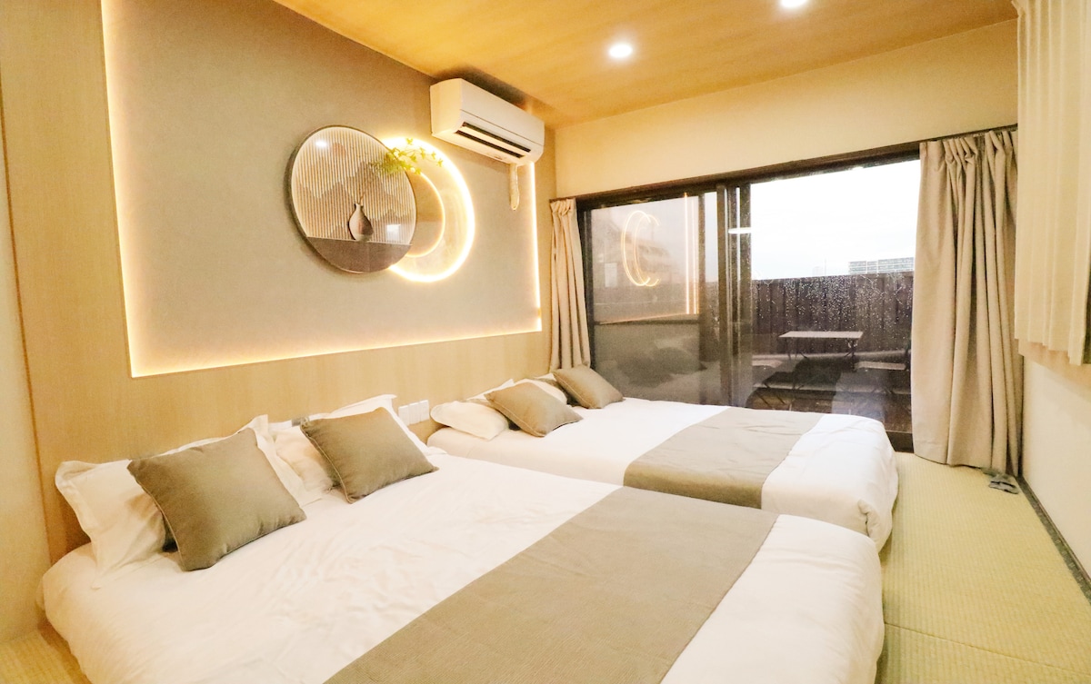 The guest room features two spacious beds with neutral-toned bedding, complemented by decorative cushions. Natural light floods the space through a large sliding door that opens to a balcony, where outdoor seating is visible. Soft, warm lighting outlines the room's modern design.