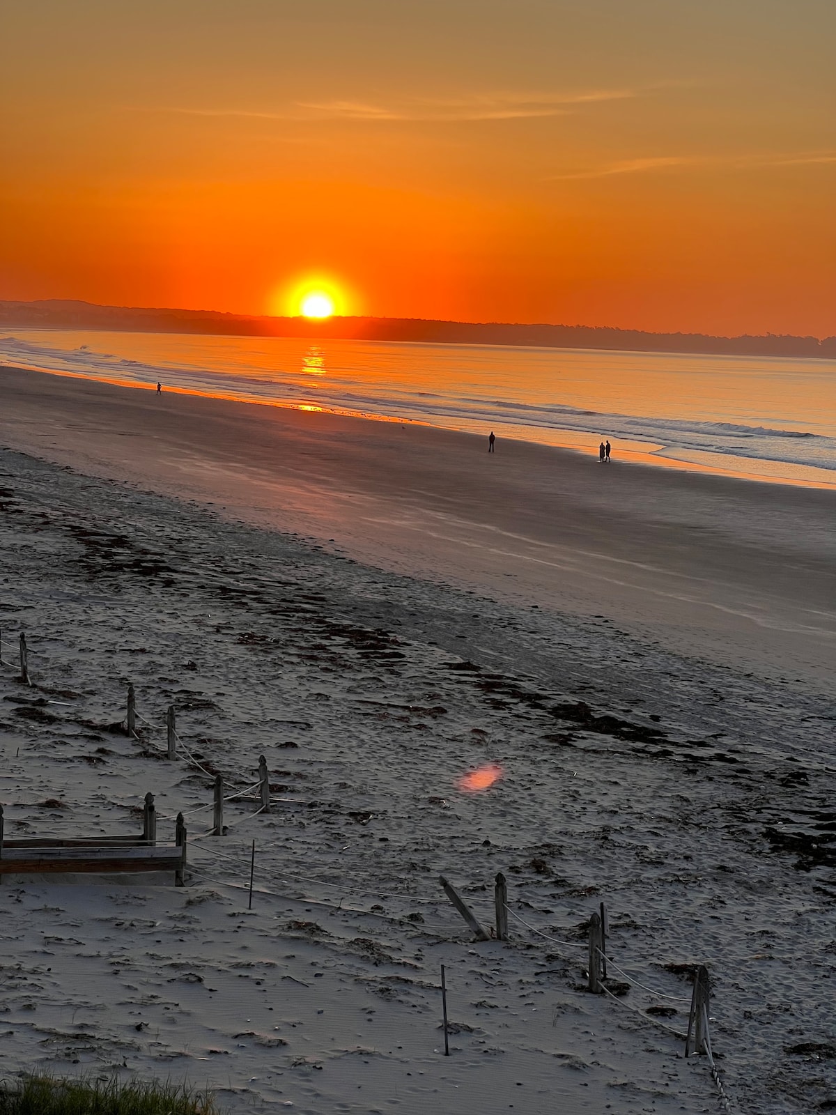 A tranquil beach scene is highlighted by a vibrant sunset casting warm orange and yellow hues over the calm waters. Silhouettes of two people walk along the shoreline, while the gentle waves lap at the sandy beach, creating a peaceful atmosphere.