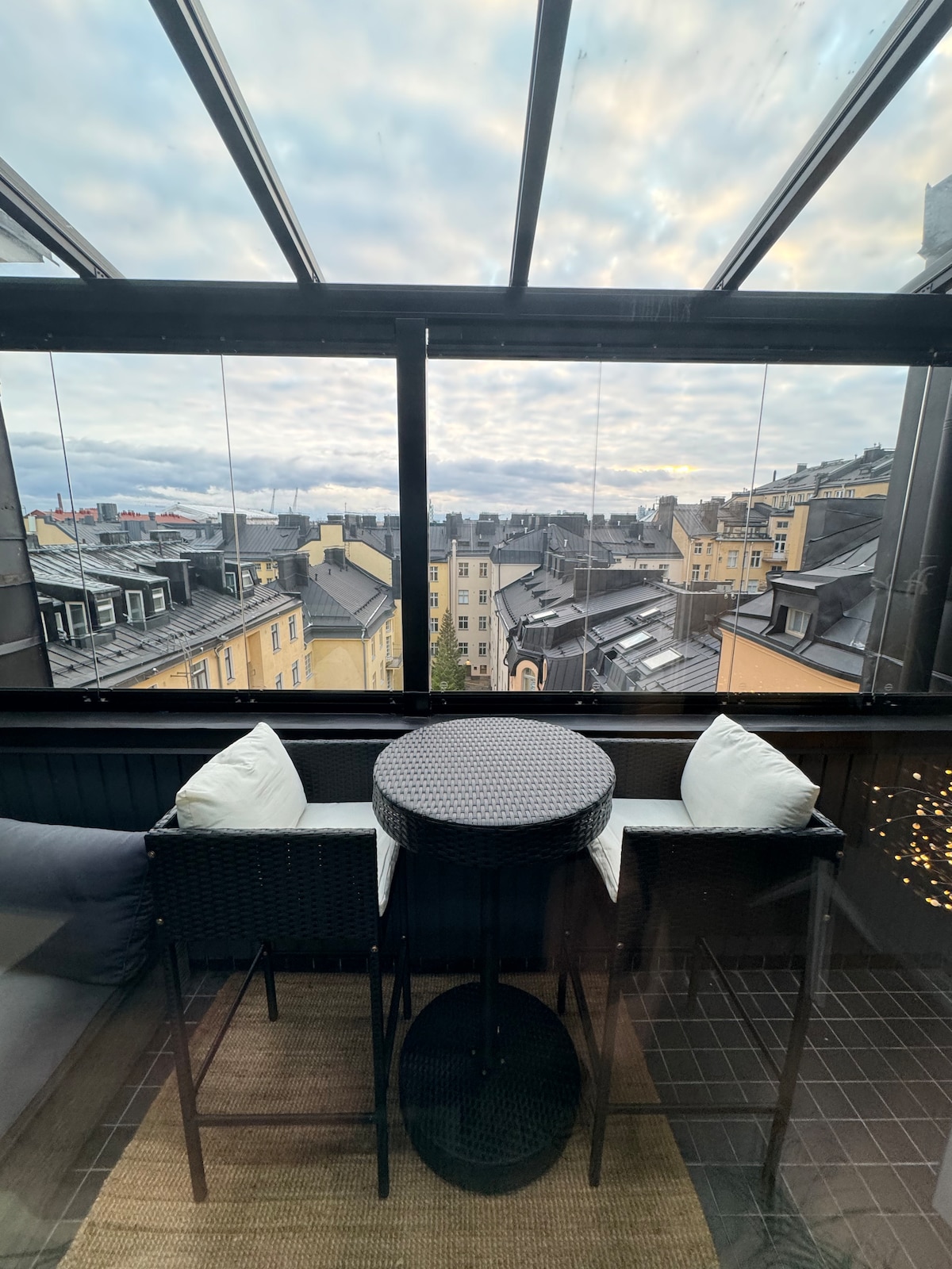 A cozy balcony is presented with a circular table and two wicker chairs, inviting relaxation. Glass panels provide an unobstructed view of the rooftops of Helsinki under a cloudy sky. The floor is covered with a natural fiber rug, enhancing the outdoor experience.