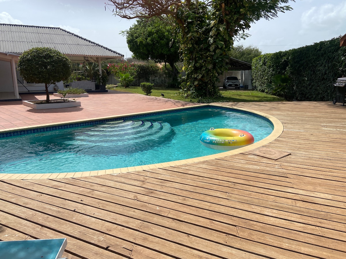 The image showcases a private swimming pool with inviting turquoise water, framed by wooden decking. A colorful inflatable float rests on the water's surface. Lush greenery is visible in the background, along with a shaded area created by the house and surrounding plants.