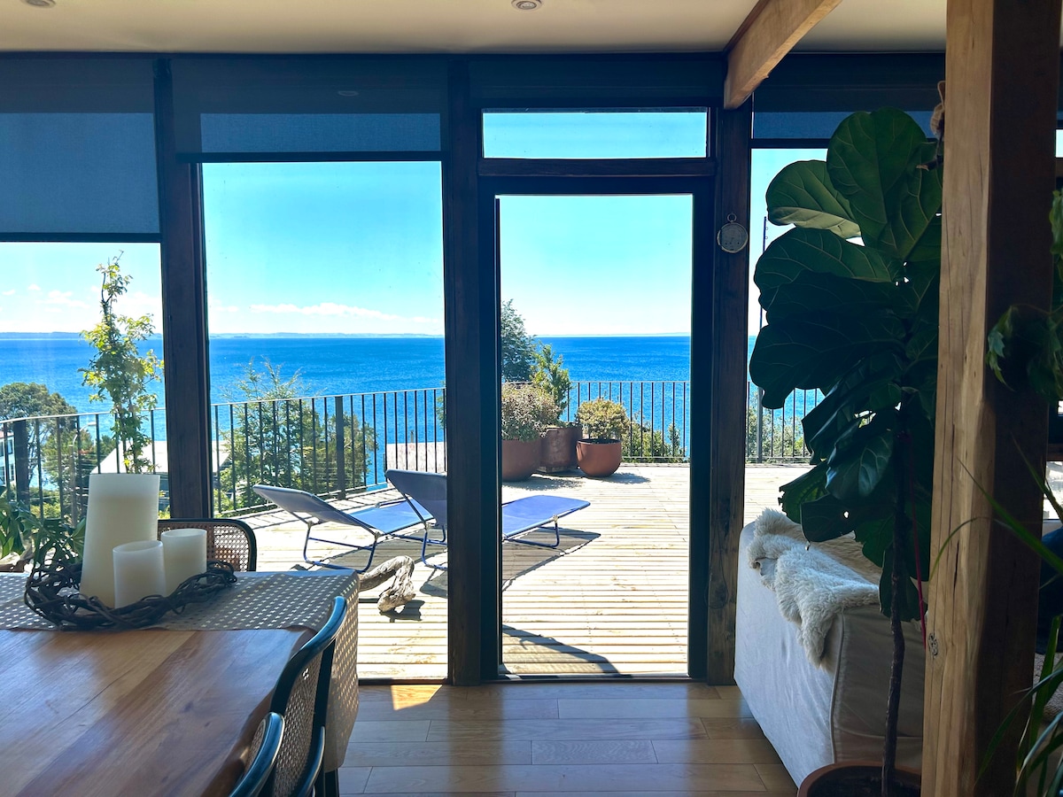 Large glass doors open to a panoramic view of the lake, allowing natural light to illuminate the space. A dining area is visible, accompanied by a cozy seating area. Outdoor furniture can be seen on the wooden deck, surrounded by greenery.