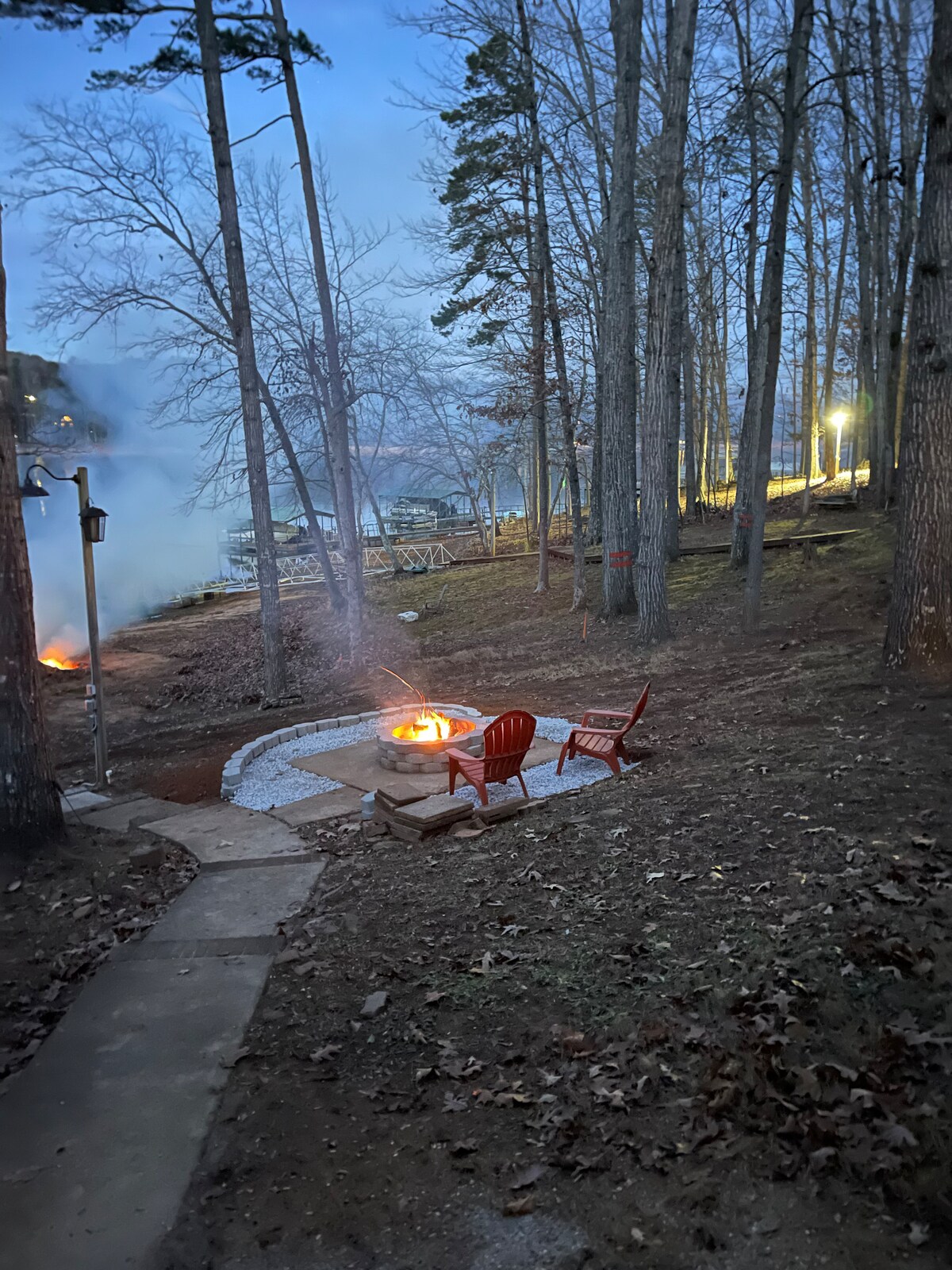 A tranquil outdoor setting features a fire pit surrounded by two red adirondack chairs, nestled among tall trees. Soft evening light casts a warm glow, and a nearby pathway leads to the lake, where a dock is visible in the distance.