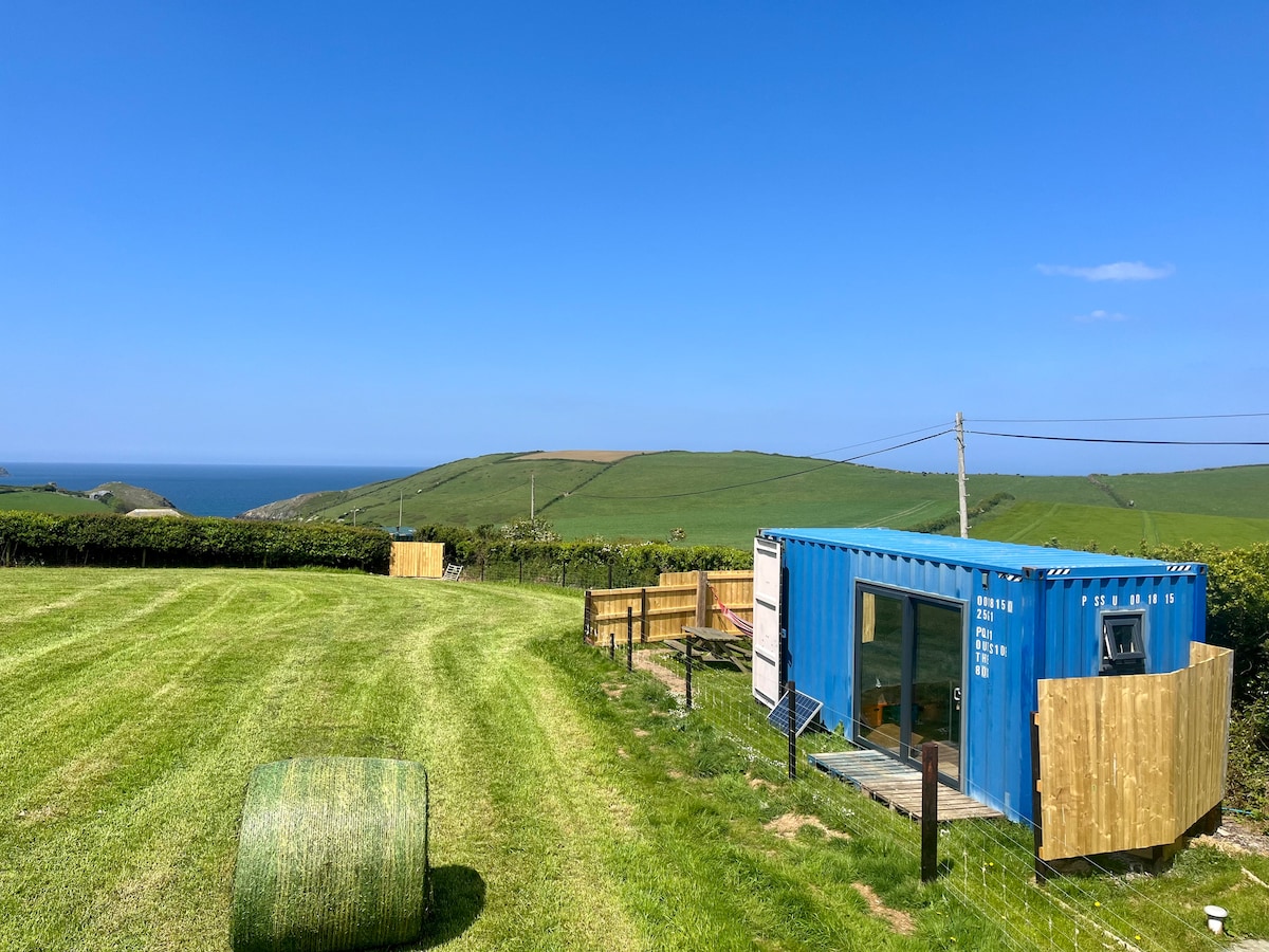 A bright blue container cabin is positioned on a grassy expanse overlooking the sea. The landscape features rolling green hills under a clear blue sky. Fencing surrounds the cabin, offering a sense of privacy, while a round hay bale can be seen in the foreground.