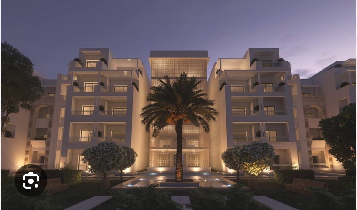 The residential building presents a modern architectural design, illuminated in soft light at dusk. Lush palm trees and manicured greenery frame a central water feature, creating a serene and inviting entrance. Spacious balconies provide aesthetic appeal to the structure.