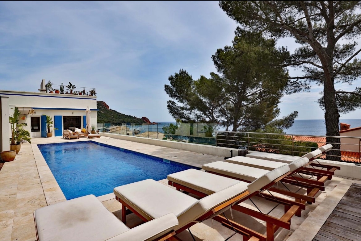 A swimming pool is framed by several sun loungers arranged neatly along the terrace. Lush greenery and trees provide a natural backdrop, while the ocean is visible in the distance under a clear sky. The villa's exterior features large sliding doors that open onto the pool area.