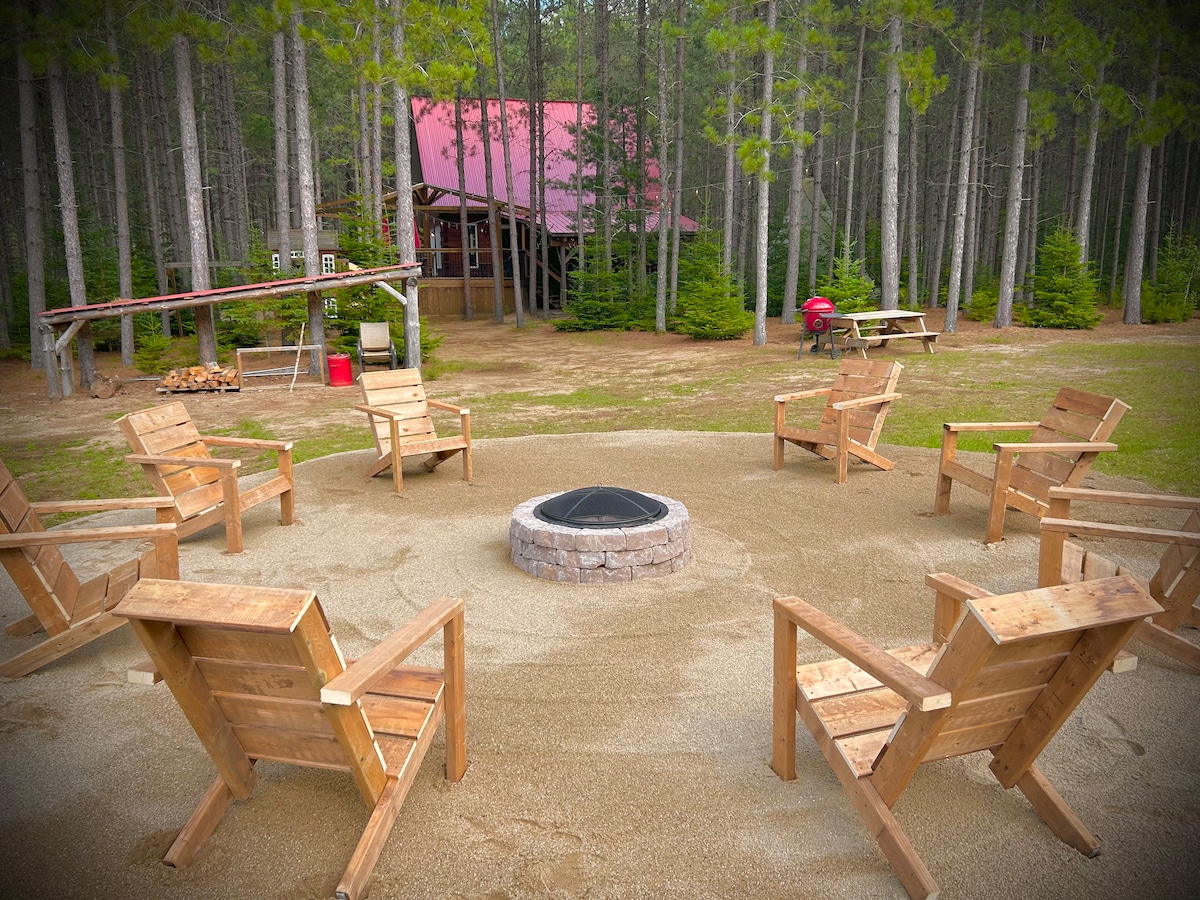 A fire pit is centered within an arrangement of eight wooden chairs, set on a sandy area surrounded by tall pine trees. A rustic wooden structure is visible in the background, alongside a picnic table and firewood storage.