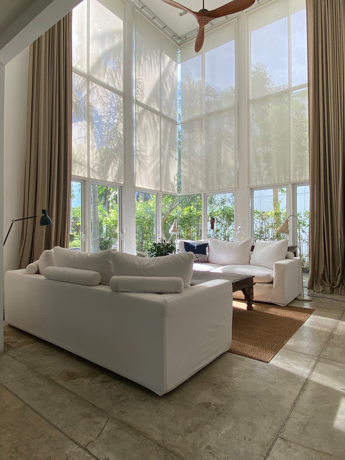 A light-filled living area is highlighted by large windows, offering views of greenery. A white sofa is arranged around a wooden coffee table, complemented by a textured area rug. Tall curtains and a ceiling fan add to the airy and spacious ambiance.