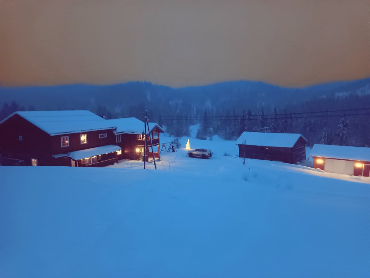 A serene winter landscape is depicted, featuring snow-covered ground and distant mountains under an evening sky. The two-story farmhouse and additional structures are softly illuminated, highlighting their rustic charm. A swing set is visible in the foreground, adding to the peaceful ambience of the property.
