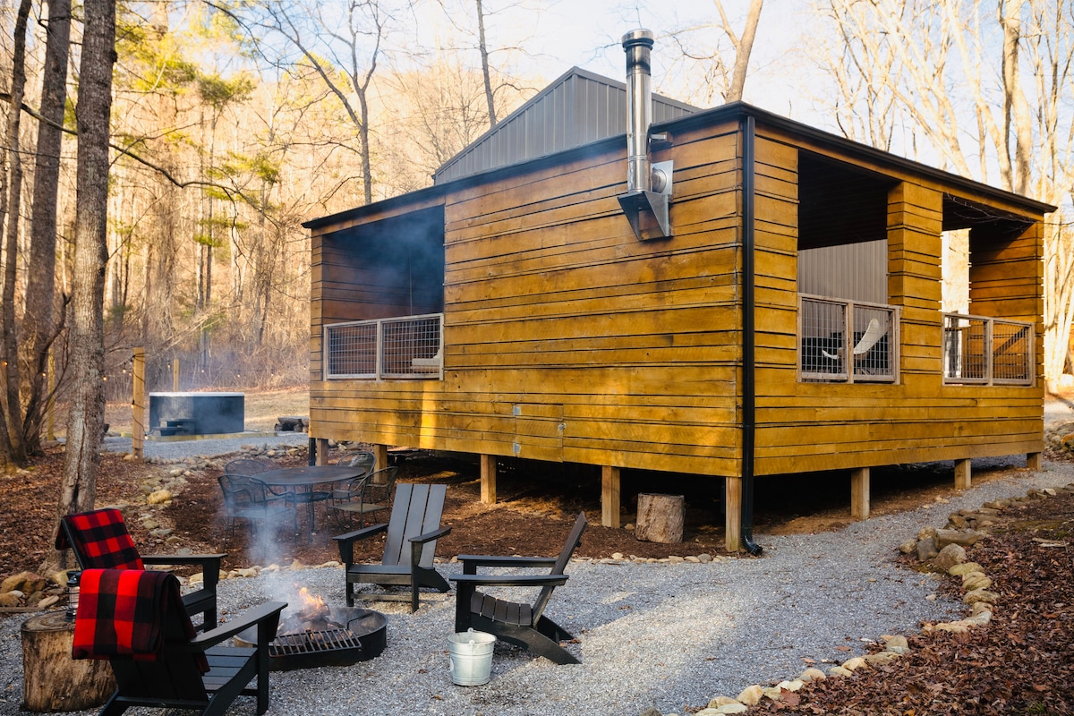 A modern cabin constructed from warm-toned wood is set amidst trees, featuring a raised porch. A fire pit with several adirondack chairs is positioned in the foreground, while a hot tub and gravel pathway are visible nearby.