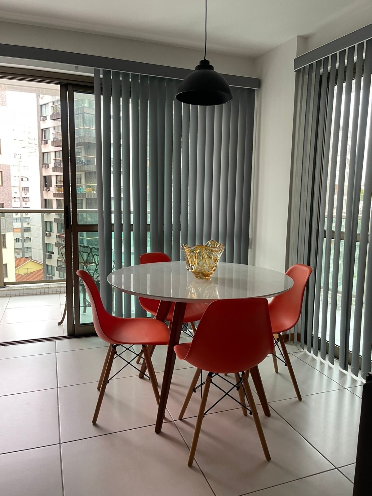An inviting dining area features a round white table surrounded by four orange chairs. Large windows with vertical blinds allow natural light to fill the space, offering a view of the city. A stylish pendant light hangs overhead, creating a modern feel.
