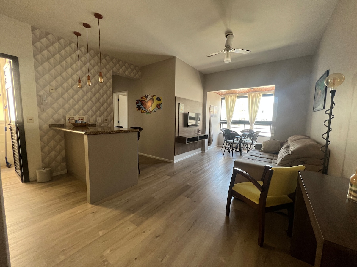 An open living area features a seamless flow between the kitchen and lounge. Light wood flooring complements the neutral walls. A comfortable sofa and an armchair provide seating. Large windows allow natural light to illuminate the space, offering a view of the outdoor balcony.