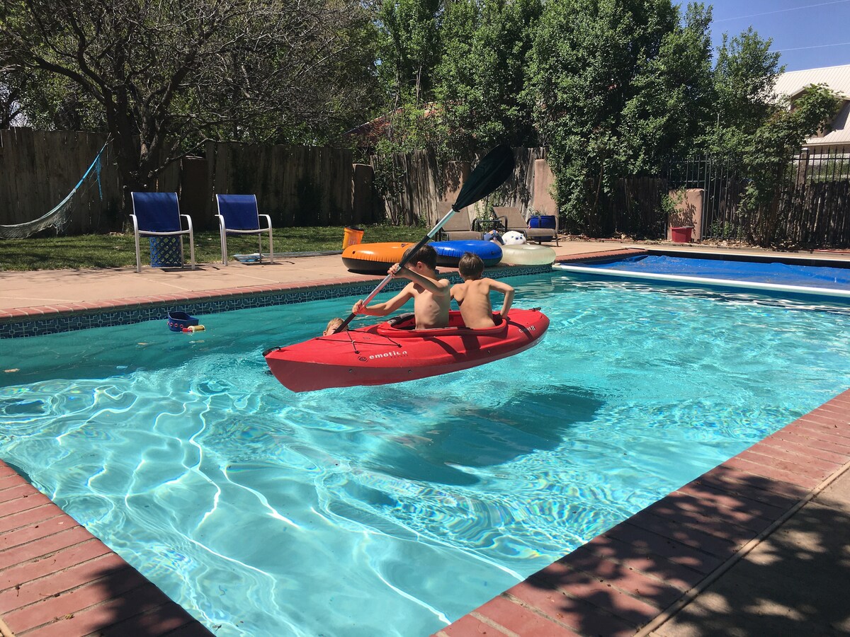 Two individuals are seen paddling a red kayak in a private pool surrounded by a brick patio. Lounge chairs are positioned nearby, with vegetation creating a natural backdrop. The pool's clear water reflects sunlight, providing a relaxing atmosphere.