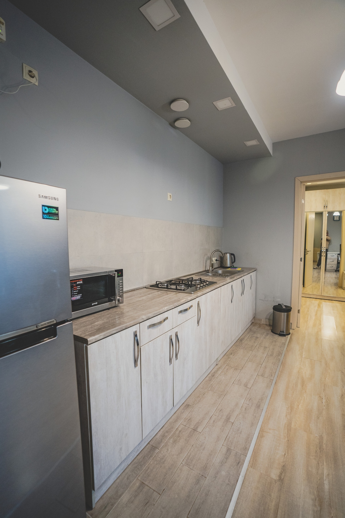 A modern kitchen featuring sleek cabinetry and a clean countertop. Essential appliances, including a refrigerator, microwave, and stovetop, are visible. The space is well-lit with ceiling lights, and a doorway can be seen leading to another area of the apartment.