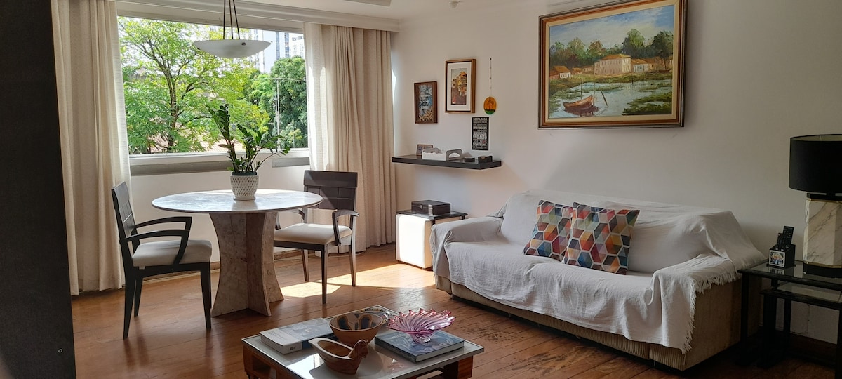 A spacious living area is presented with a round dining table surrounded by two chairs. A light-colored sofa, draped with a cover, is adorned with colorful cushions. Natural light filters through large windows, illuminating the wooden flooring and artwork on the walls.