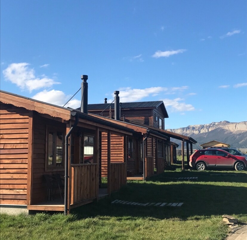 A cluster of independent cabins is presented amidst a grassy area. Each cabin features wooden exteriors, complemented by sloped roofs and chimney stacks. A vehicle is parked nearby, with scenic mountains visible in the background, enhancing the natural surroundings.