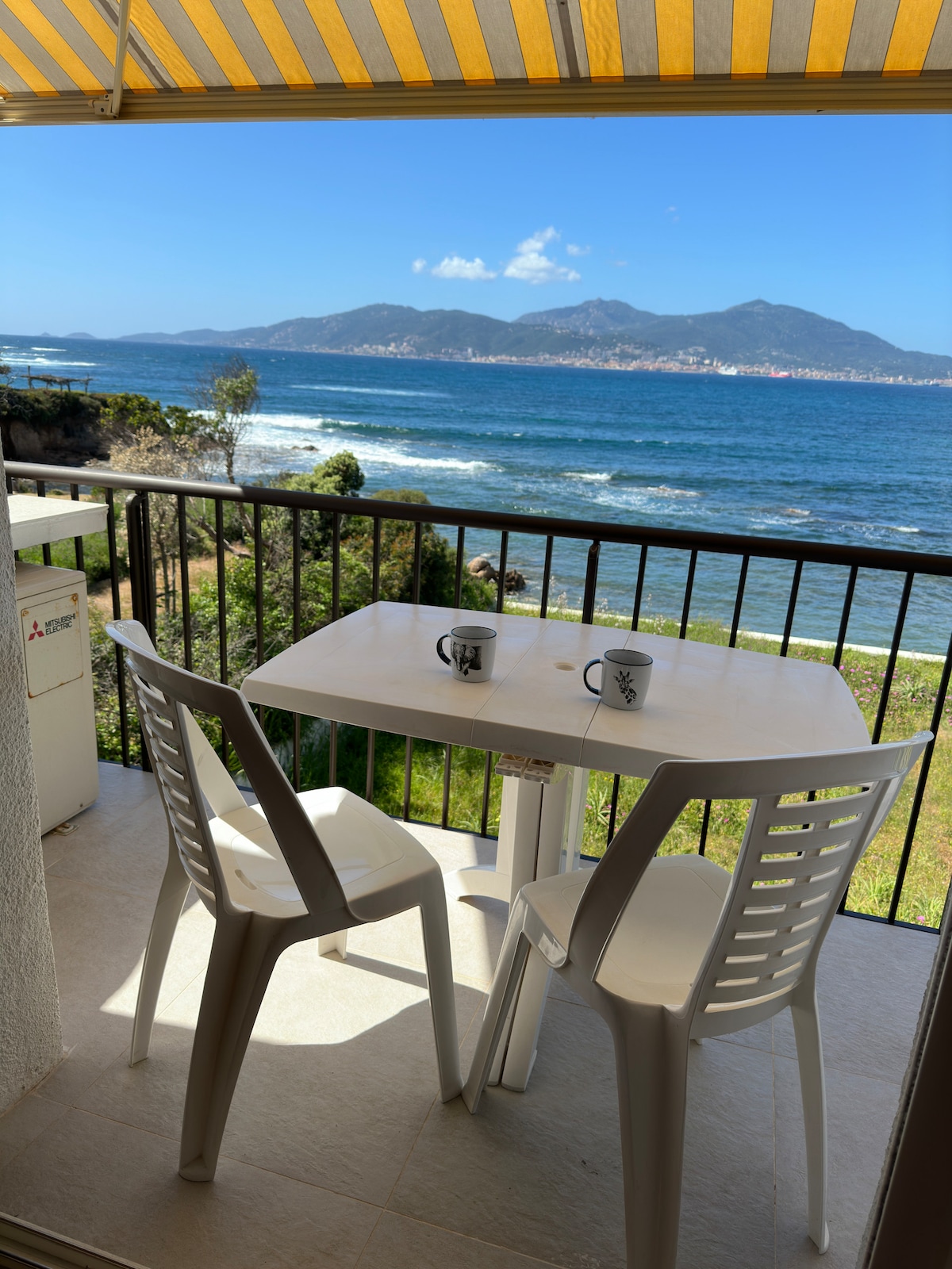 A balcony with a sea view features a white table and two plastic chairs, providing a spot for relaxation. The scene includes distant mountains and rolling waves, with sunlight illuminating the area. Yellow and white striped awnings provide shade.