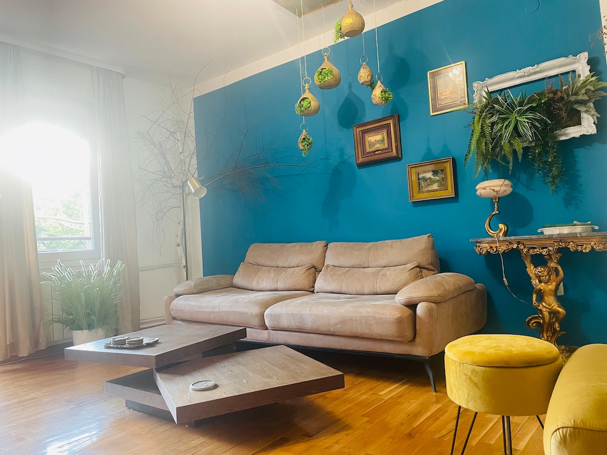 A cozy living room is presented with a light brown sofa set against a vibrant blue wall. Decorative elements include framed art and greenery. A unique coffee table is positioned in the center, with a yellow stool nearby, and natural light filters through the window.