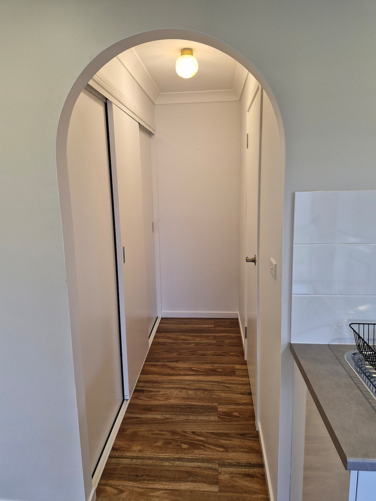 A hallway is displayed, featuring smooth wooden flooring and a simple archway. Light from a ceiling bulb illuminates the space, which includes two sliding doors along one side, leading to storage areas and a continuation toward the kitchen at the end.