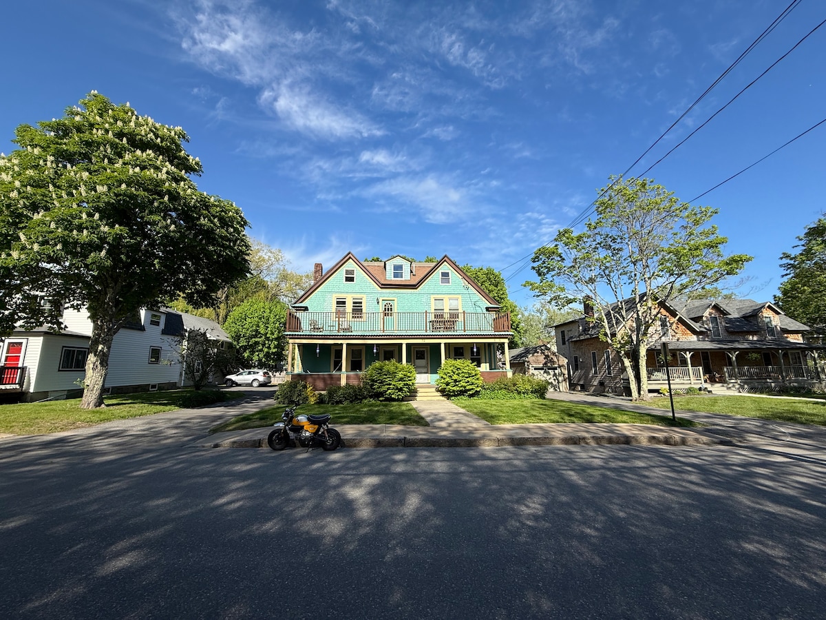 A two-story structure is displayed with a light blue exterior and multiple balconies. The front garden is landscaped with greenery, and a walkway leads to the entrance. Nearby trees provide shade, and a motorcycle is parked along the street, adding a casual element to the scene.