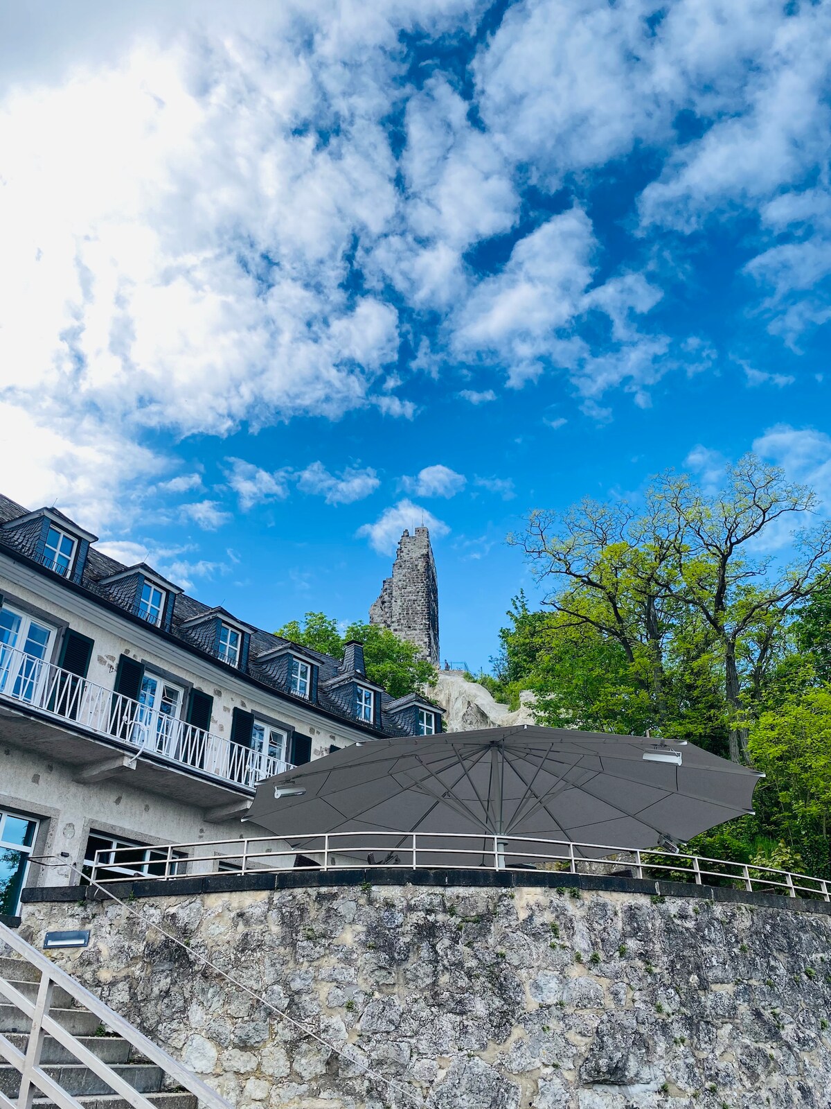 A two-story building is shown with multiple windows and balconies, set against a backdrop of a tall stone structure. A large umbrella provides shade on the terrace area, while greenery is visible nearby under a bright blue sky with scattered clouds.