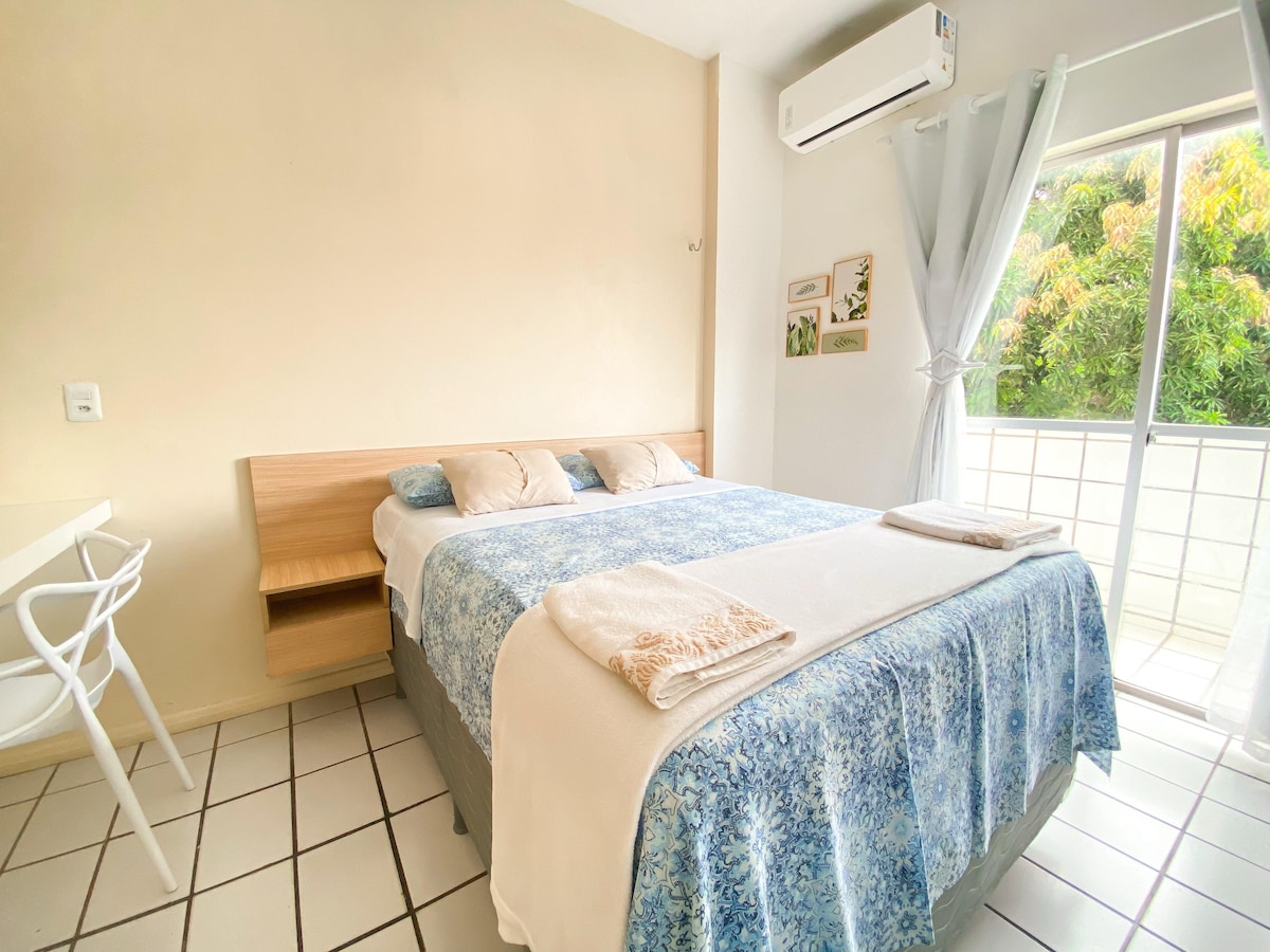 A spacious bedroom is presented with a large bed featuring a blue-patterned blanket and white linens. Two pillows rest on the bed, and a small table with a chair is positioned nearby. Natural light enters through a glass door leading to a balcony, surrounded by greenery.