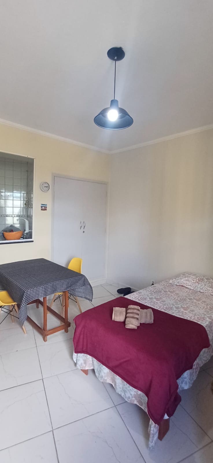 A cozy space features a double bed covered with a maroon blanket and rolled towels atop. A small wooden table with four yellow chairs is positioned nearby, alongside a window with a glimpse of the kitchen. Soft lighting emanates from a pendant lamp above.