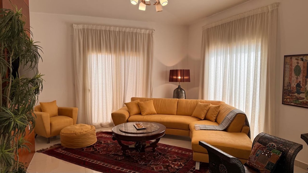 A spacious living area features a contemporary yellow sectional sofa and an accent chair, complemented by a round coffee table set on an intricate oriental rug. Natural light filters through sheer drapes, adding warmth to the room. Decorative wall art enhances the inviting ambiance.