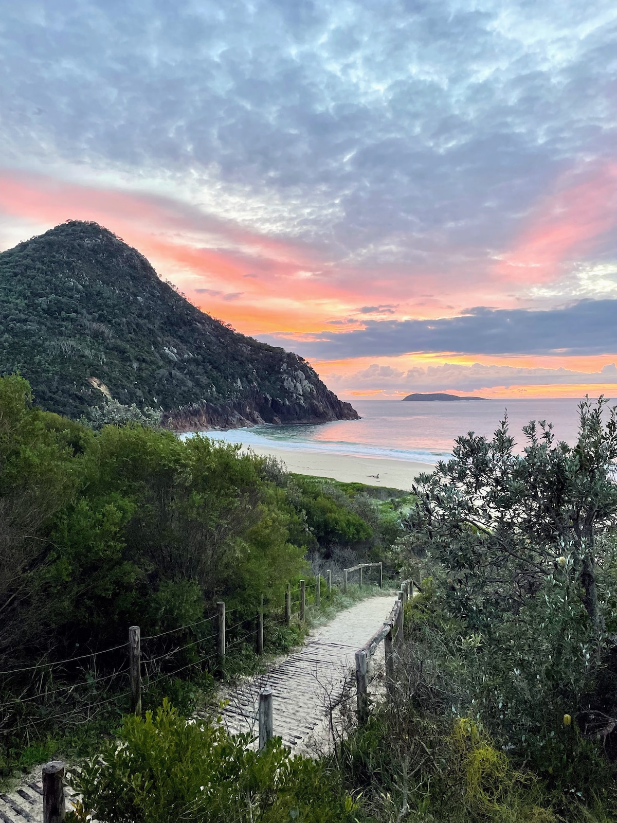 A winding pathway leads through lush greenery towards a tranquil beach, framed by a dramatic mountain. The sky displays a blend of soft pastel colors at sunset, reflecting off the gentle waves of the ocean in the background.