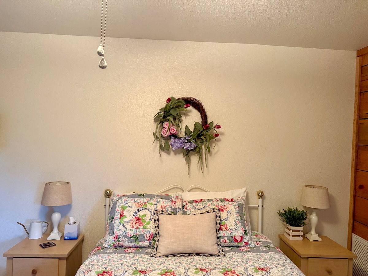 A queen bed is adorned with a floral-patterned quilt and decorative pillows. A wooden headboard and two matching bedside lamps provide soft lighting. A wreath made of flowers and greenery is displayed above the bed, enhancing the cozy ambiance of the room.