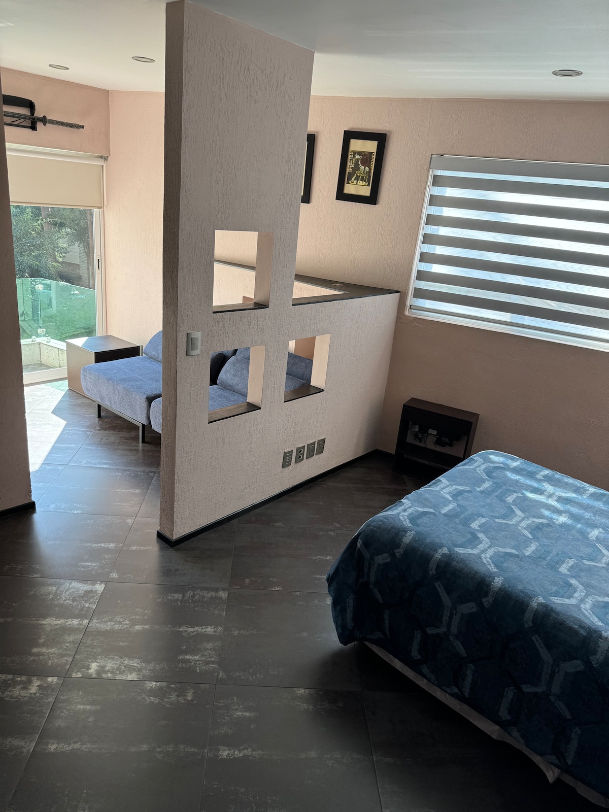 A spacious suite features a cozy bed with a blue patterned blanket, alongside a partition that allows for an open flow to the living area. A comfortable seating space is visible near large sliding glass doors, letting in ample natural light.