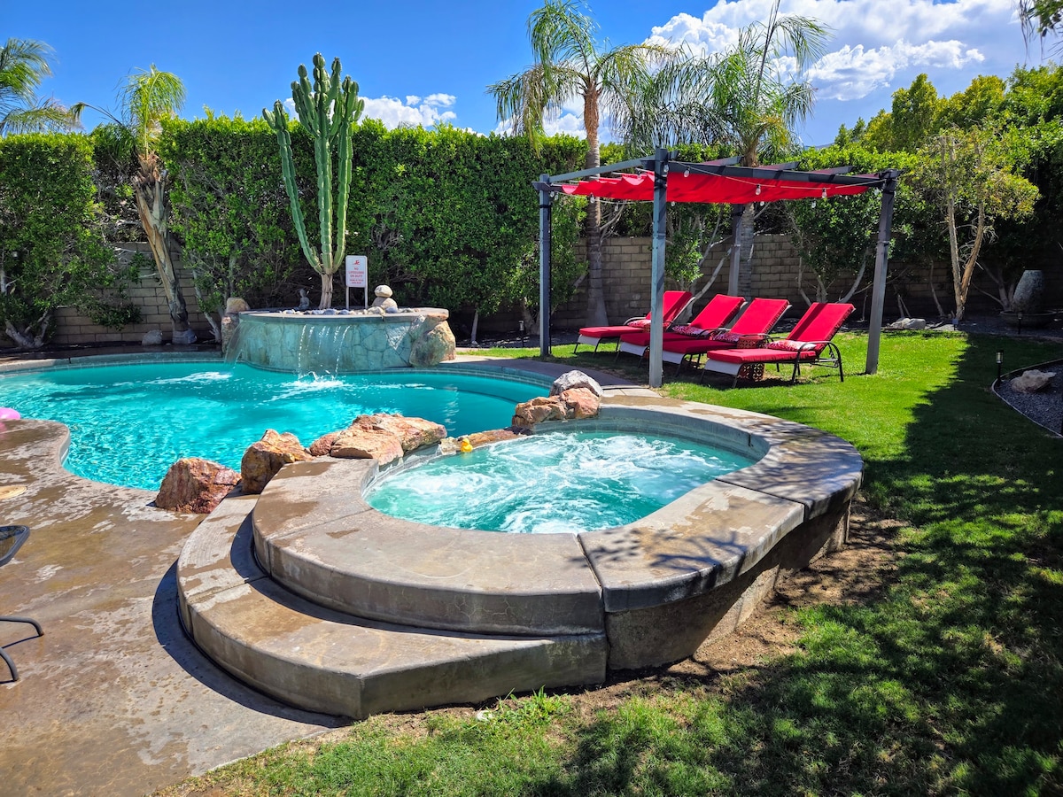 A serene private backyard showcases a saltwater pool and spa surrounded by lush greenery. A waterfall feature adds tranquility to the scene, while red lounge chairs provide comfortable seating. Sunlight reflects off the water, enhancing the inviting atmosphere and natural beauty of the space.