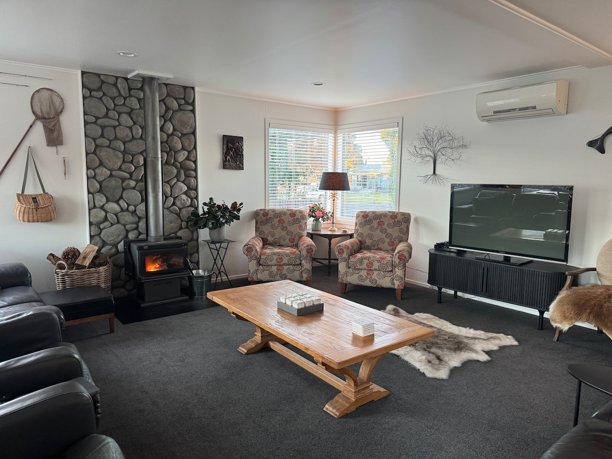 A spacious living area features two floral armchairs positioned across from a modern television. A wooden coffee table centers the room, complemented by a cozy area rug. Natural light streams through large corner windows, while a wood-burning stove provides warmth.