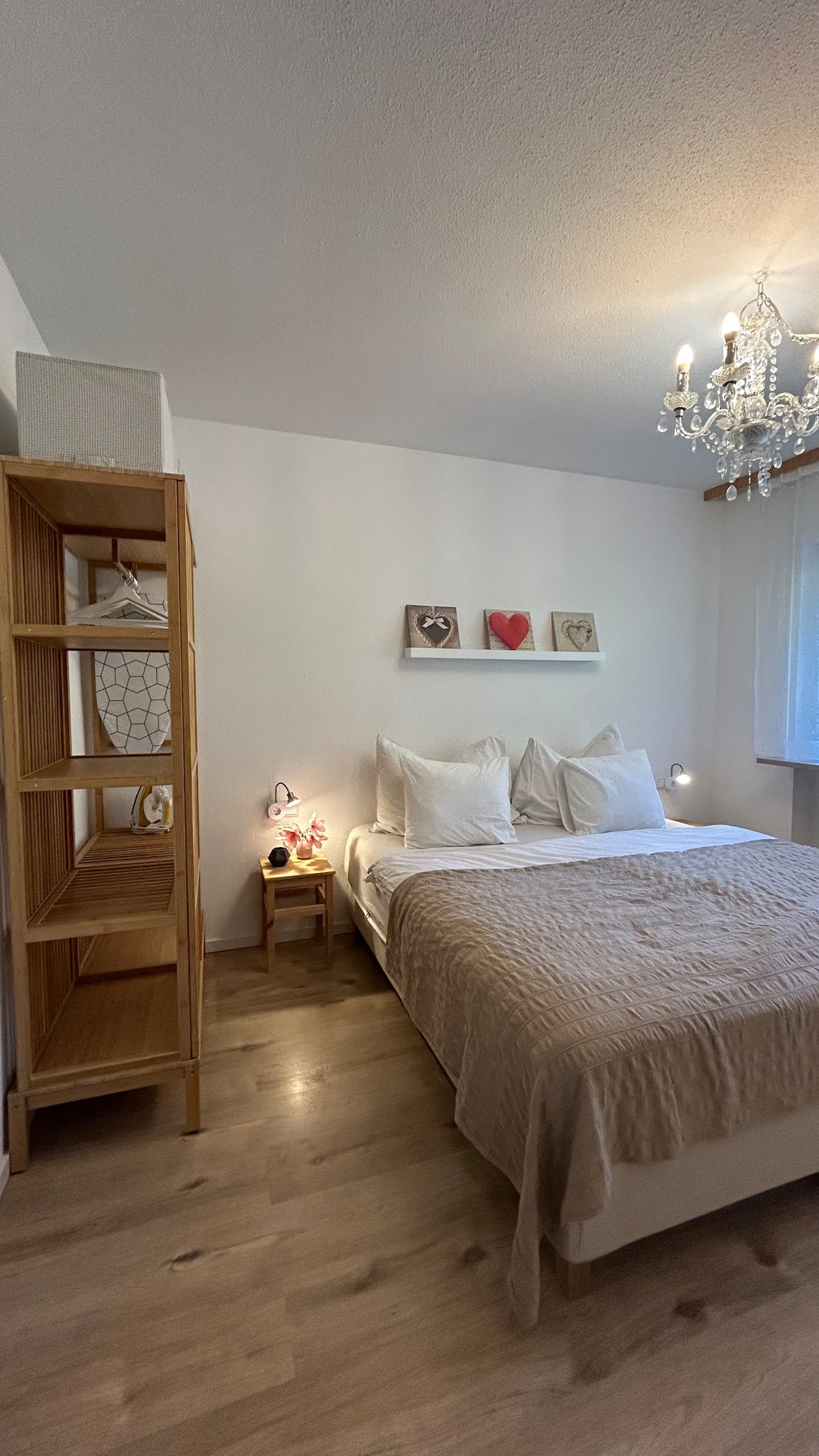 The bedroom features a spacious bed with white bedding and decorative pillows. A wooden shelf displaying framed heart art enhances the wall. A charming chandelier hangs from the ceiling, while a bedside table and a bookshelf provide functional storage.