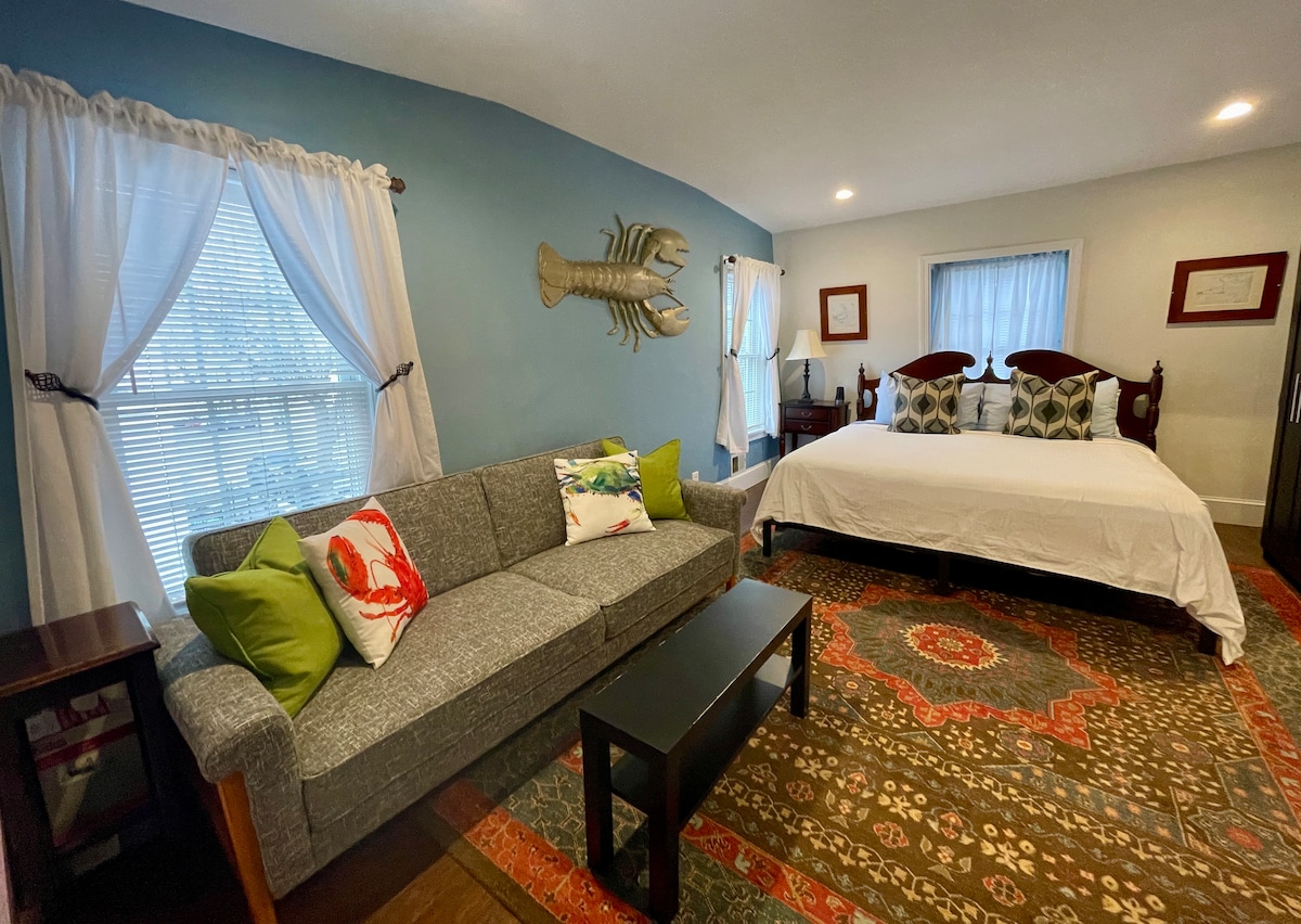 A spacious suite features a king EuroTop bed adorned with decorative pillows. A gray sofa with colorful accent pillows and a dark coffee table complement the room. Natural light enters through two windows, with light-colored curtains framing them. An eclectic rug adds warmth to the wooden floor.