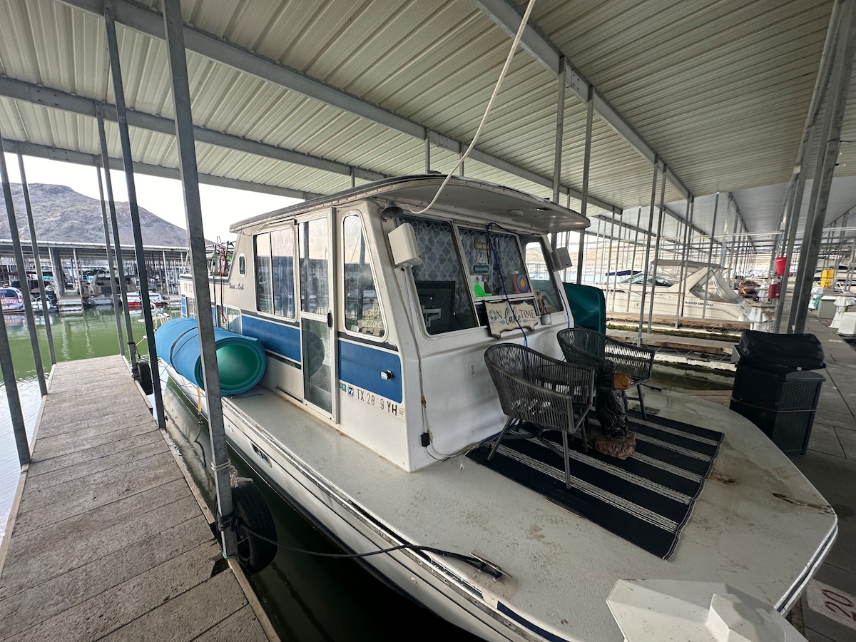 A houseboat is stationed in a covered marina, featuring a spacious deck with black and white striped rug. Two chairs are positioned at the helm, with a clear view beyond the protective railing. The surrounding area showcases other boats and the calm water.