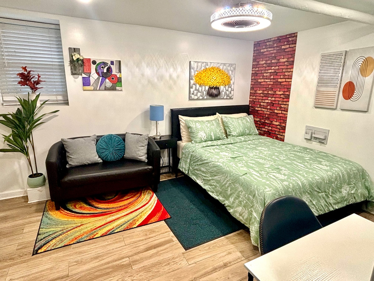 A cozy sleeping area features a spacious bed covered with a green patterned duvet. A dark couch with a decorative pillow complements the room, which includes a vibrant area rug. Artwork adorns the walls, with a striking brick accent providing visual interest.