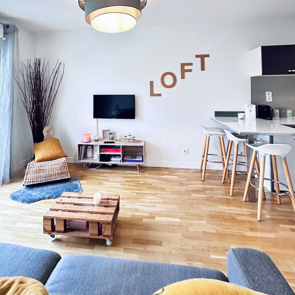 The open living space features wooden flooring and a comfortable seating area with a gray sofa. A rustic coffee table on wheels sits in front of the seating. A wall-mounted television is visible alongside modern bar stools and an inviting nook with natural decor elements.