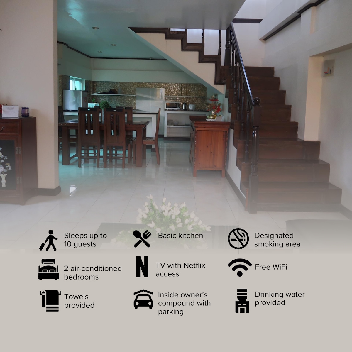 The open-plan living area features a dining table with chairs, positioned near the kitchen, visible in the background. A staircase leads to the second floor, where the air-conditioned bedrooms are located. Light-colored tiles enhance the spaciousness of the area.