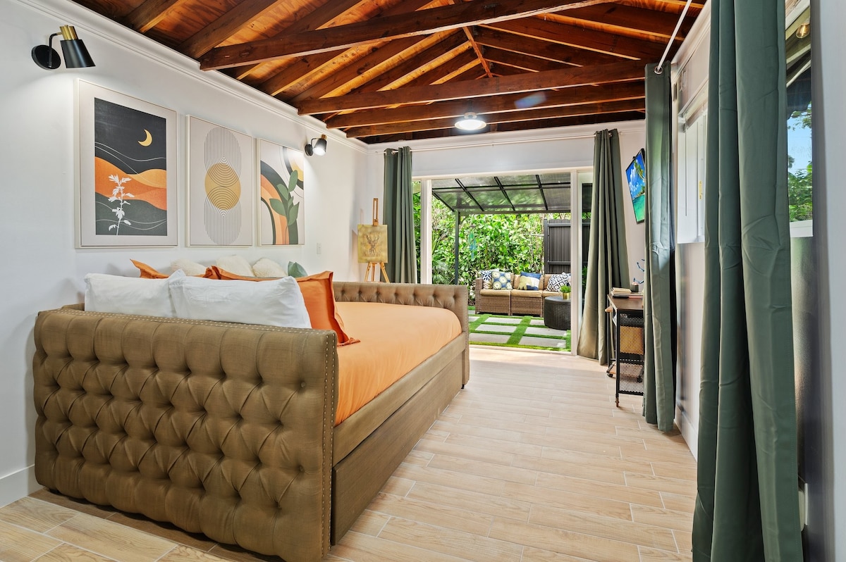 A well-appointed sleeping area features a tufted bed covered with orange linen and decorative pillows. Artwork is displayed above the bed, and soft natural light enters through large windows, accentuating the wooden ceiling beams and stone-tiled floor.