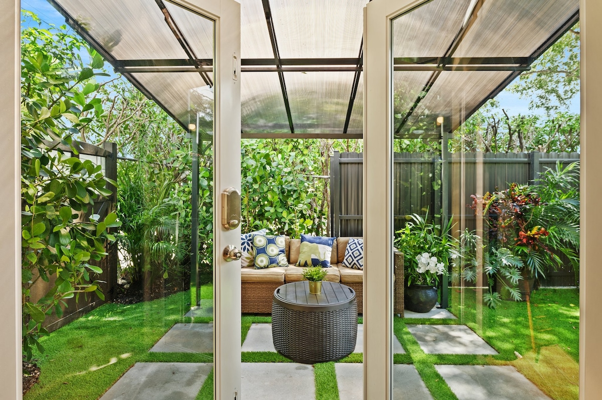 A private backyard is framed by double glass doors, showcasing a furnished outdoor space. A comfortable sofa with decorative cushions sits next to a round table, surrounded by lush greenery and vibrant flower arrangements. Soft grass and stepping stones create a serene atmosphere.