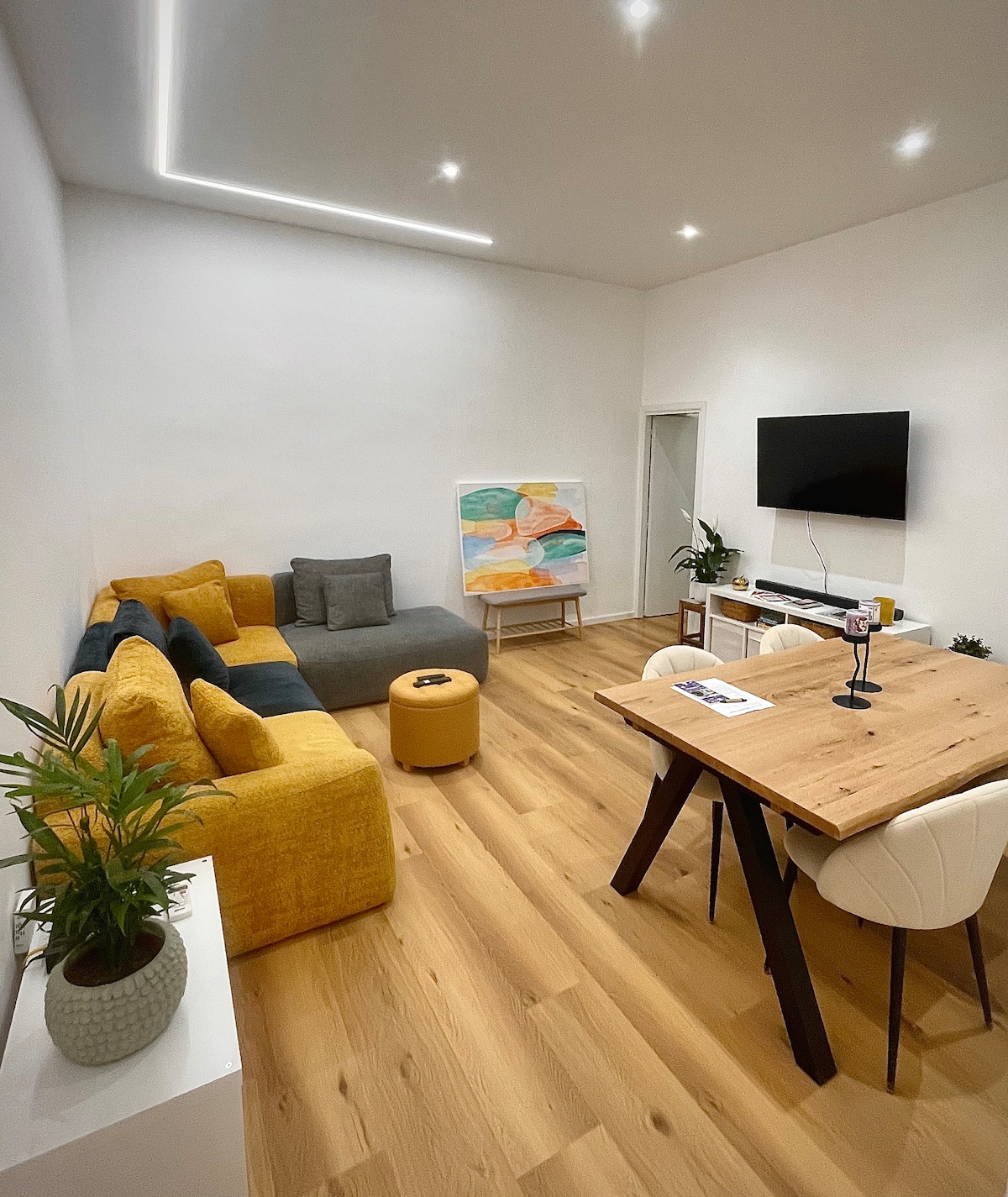 A modern living area features a sectional sofa in shades of gray and yellow, complemented by a round ottoman. A wooden dining table with light-colored chairs occupies the space, while a vibrant piece of artwork stands against the wall. A wall-mounted TV and several plants add to the atmosphere.