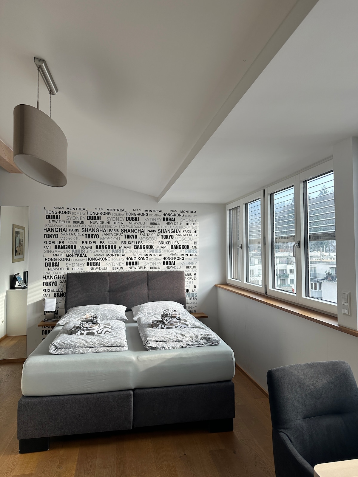 A modern sleeping area is highlighted by a cozy bed with neutral bedding, and a stylish headboard. Large windows allow natural light to fill the room, while a comfortable chair is placed nearby, creating a serene space for relaxation.