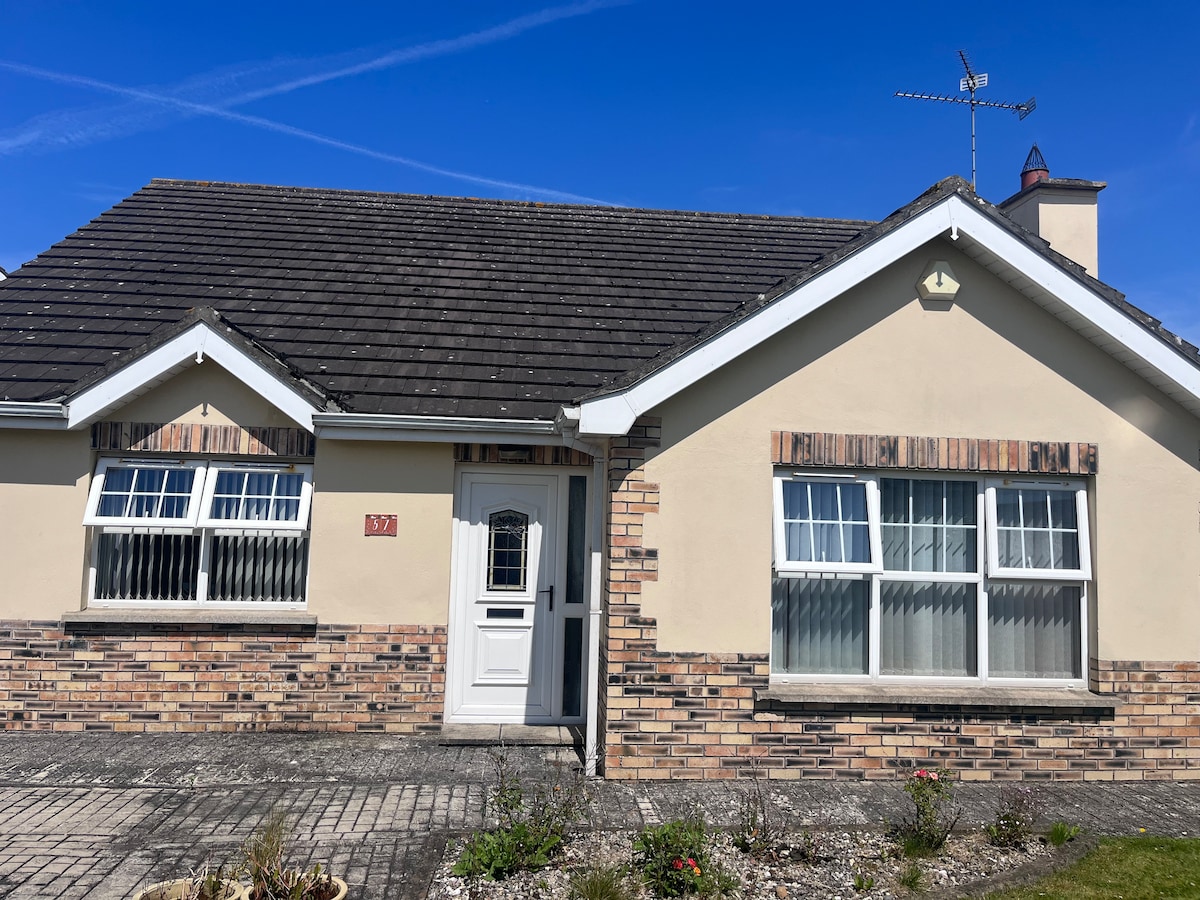 The front view of the bungalow is displayed, showcasing a brick facade with a softly sloping roof. Two large windows flank the entrance door, providing natural light to the interior. A paved area and garden beds are visible in front, offering a well-kept outdoor space.