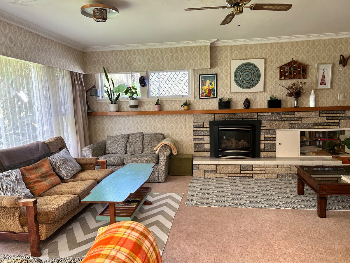 A living room features a combination of comfortable seating, including a patterned couch and armchairs. A coffee table sits on a geometric rug, while a stone fireplace adds a focal point. Natural light flows through large windows adorned with sheer curtains, showcasing indoor plants.
