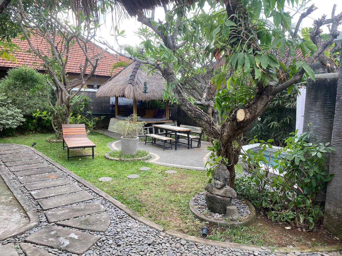 A serene garden space features a thatched pavilion surrounded by lush greenery. Comfortable lounge chairs are positioned for relaxation, while a stone path leads through the area. A small statue is visible amongst the plants, contributing to the tranquil atmosphere.