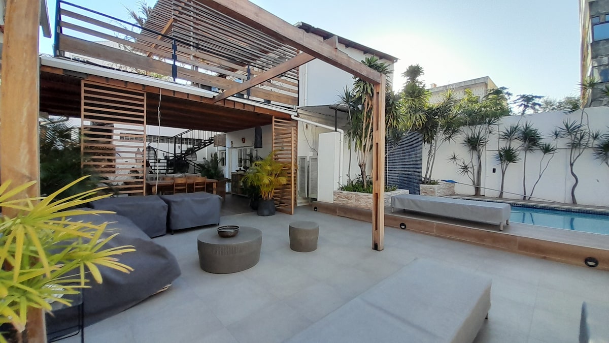 A serene outdoor deck area is presented, featuring comfortable seating arrangements with soft cushions. Lush greenery is incorporated throughout, alongside a swimming pool visible in the background. The space is designed for relaxation, with natural light casting gentle shadows across the smooth tiled floor.